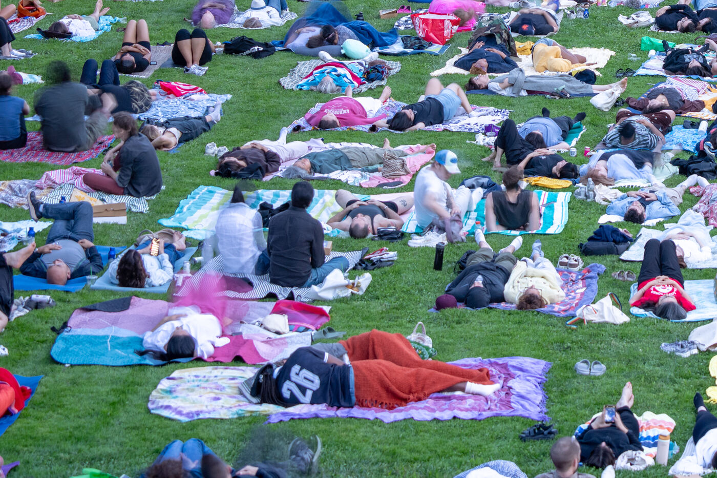 Close view of the lawn: dozens of people lying on beach towels and quilts, eyes closed, shoes off, a man asleep under a red blanket.
